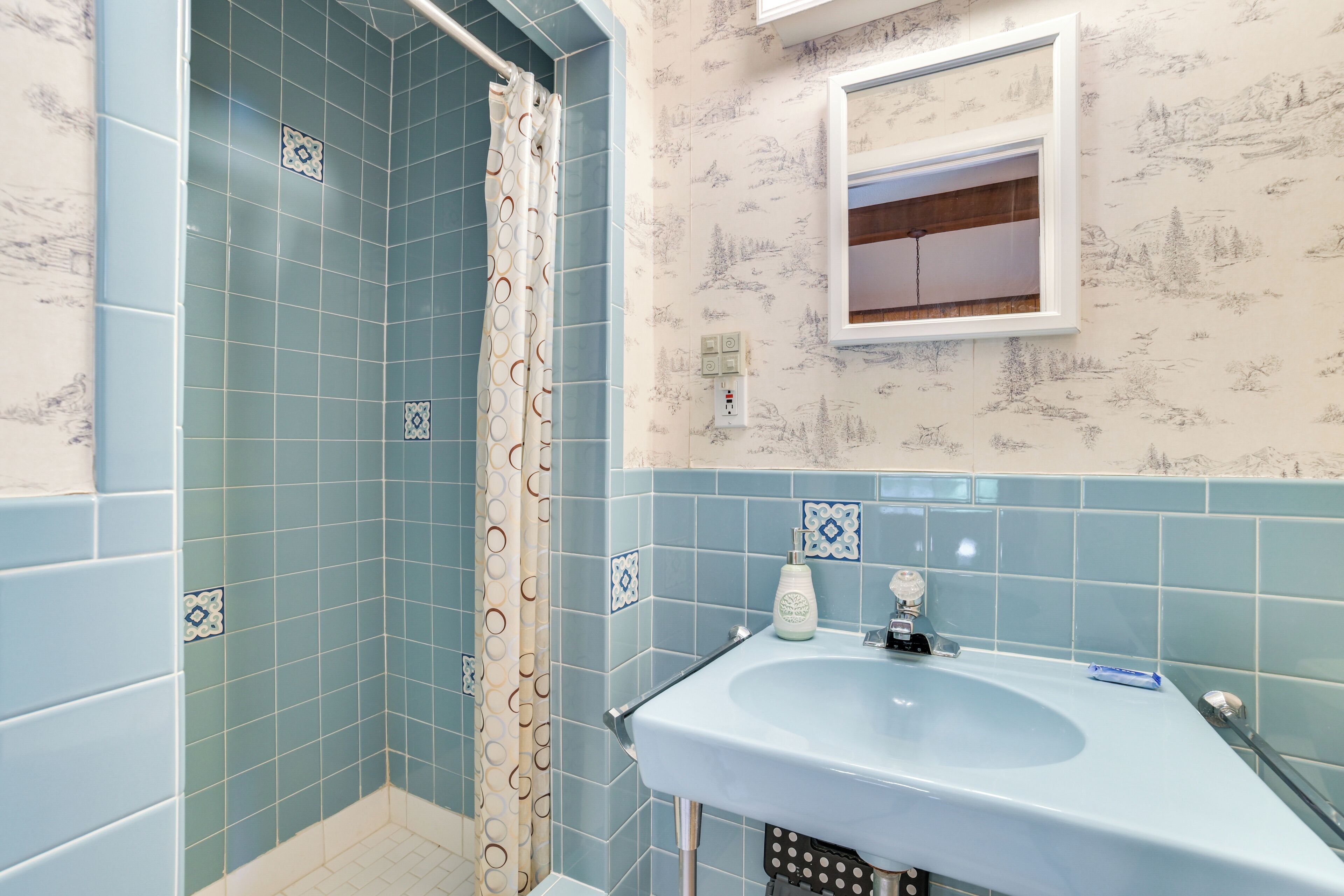 Une salle de bain avec des carreaux bleus, une douche avec un rideau à motifs, un lavabo avec un miroir au-dessus, et un mur recouvert d’un papier peint sur le thème de la nature.