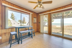 A room with a wooden interior, a ceiling fan, a table with two stools, large windows with a beach view, and a sliding glass door leading to an outdoor area.