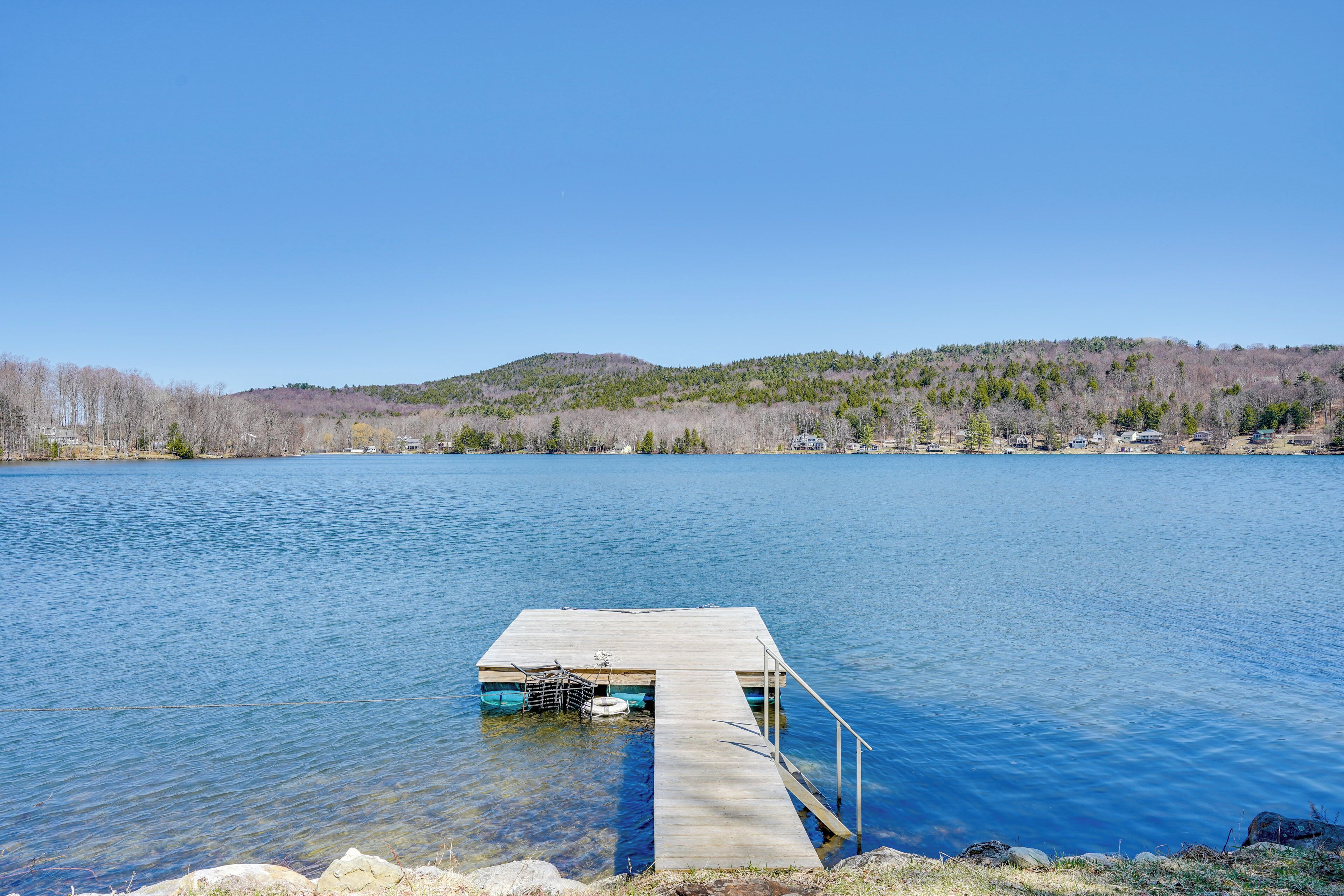 A wooden dock extending into a calm lake with a clear sky and a forested hill in the background.