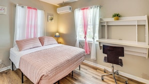 A bedroom with a bed, a ceiling fan, an air conditioner, a desk with a chair, and a window with curtains.