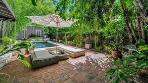 A private pool area with lounge chairs and a large umbrella, surrounded by lush greenery and a brick patio. - The Royale 2B 4B Luxury Garden Pool Yoga Room (Tampa)