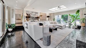 A modern living room with a sectional sofa, a coffee table, and a large window offering a view of greenery. - The Royale 2B 4B Luxury Garden Pool Yoga Room (Tampa)