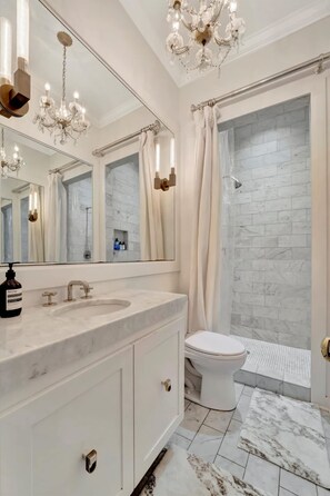 Une salle de bain avec un lavabo en marbre, des toilettes, une douche avec une porte en verre et un grand miroir agrémenté d’appliques murales.