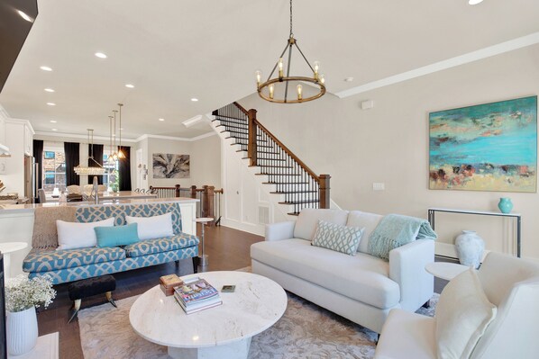 A spacious living room with a staircase, a chandelier, and a large painting on the wall.