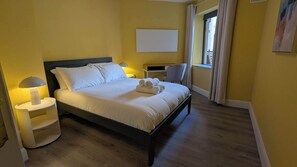 Uma cama bem arrumada com lençóis brancos, uma mesa de cabeceira com abajur e uma cadeira em um quarto com paredes amarelas e piso de madeira.