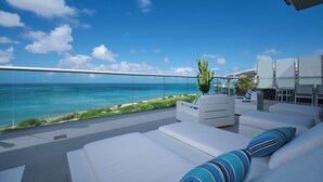 Angolo lounge esterno moderno con divani bianchi e cuscini a righe, ringhiera in vetro e vista sull'oceano.