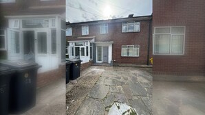 A residential area with a paved courtyard, a brick house with a white door, and two black bins with recycling labels.