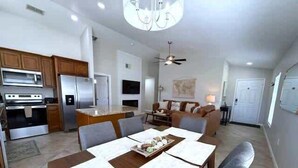 Una cocina moderna con electrodomésticos de acero inoxidable, un comedor con mesa de madera y sillas grises, y una sala de estar con sofá y ventilador de techo.