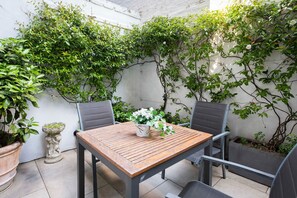 An outdoor patio with a wooden table and two chairs, surrounded by lush greenery and a white wall. - Beautiful 3-Bdr Chelsea Home with Garden patio – Steps from King’s Rd (London)
