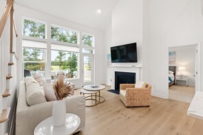 A spacious living room with a fireplace, a large TV, and a view of the outdoors through large windows.