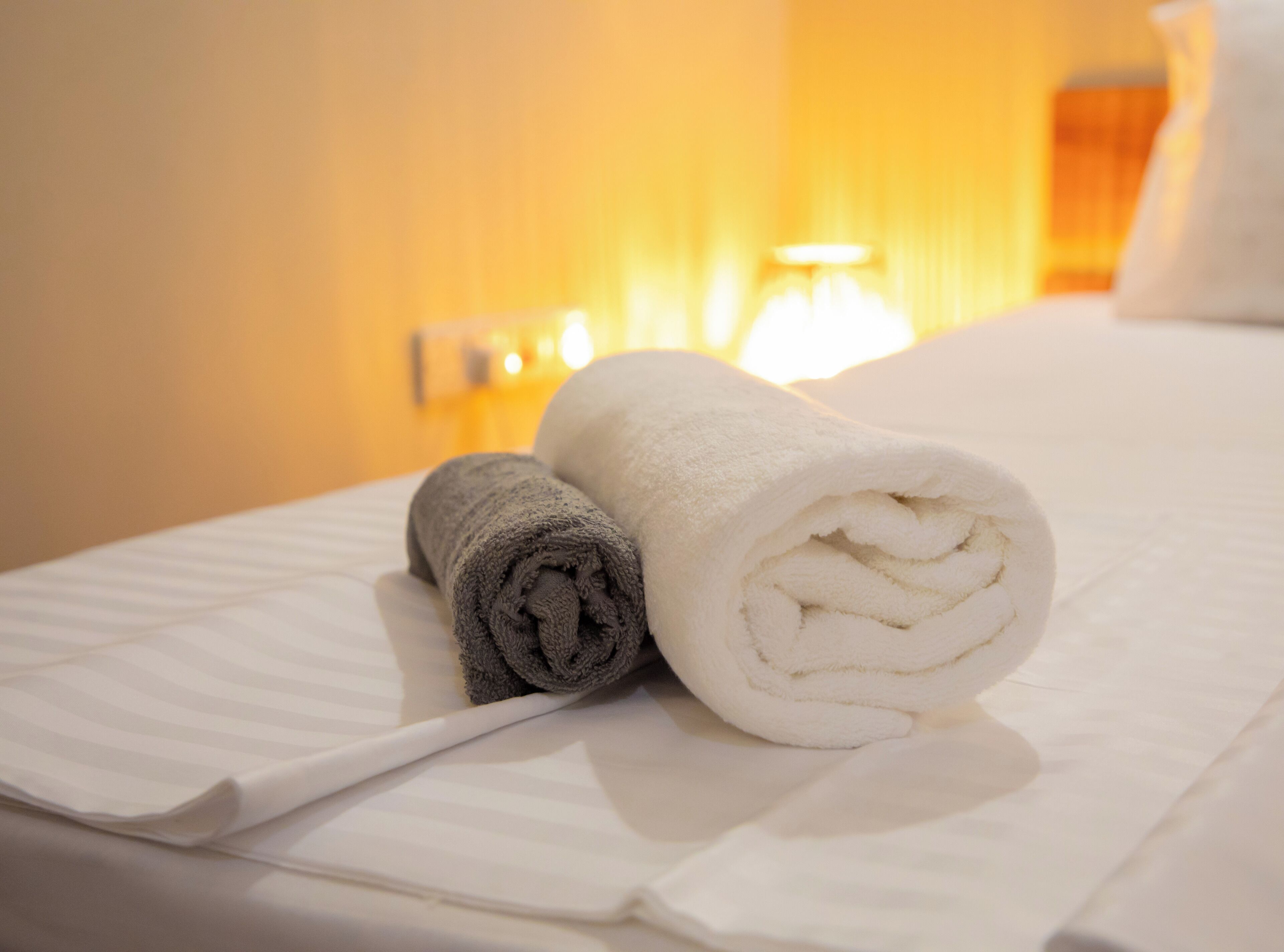 A neatly made bed with two rolled towels, one grey and one white, in a hotel room with a warm, yellow-lit ambiance.