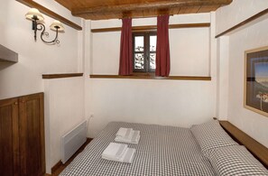 Une chambre avec une couvre-lit à carreaux, une armoire en bois, une fenêtre avec des rideaux rouges et des luminaires fixés au mur.