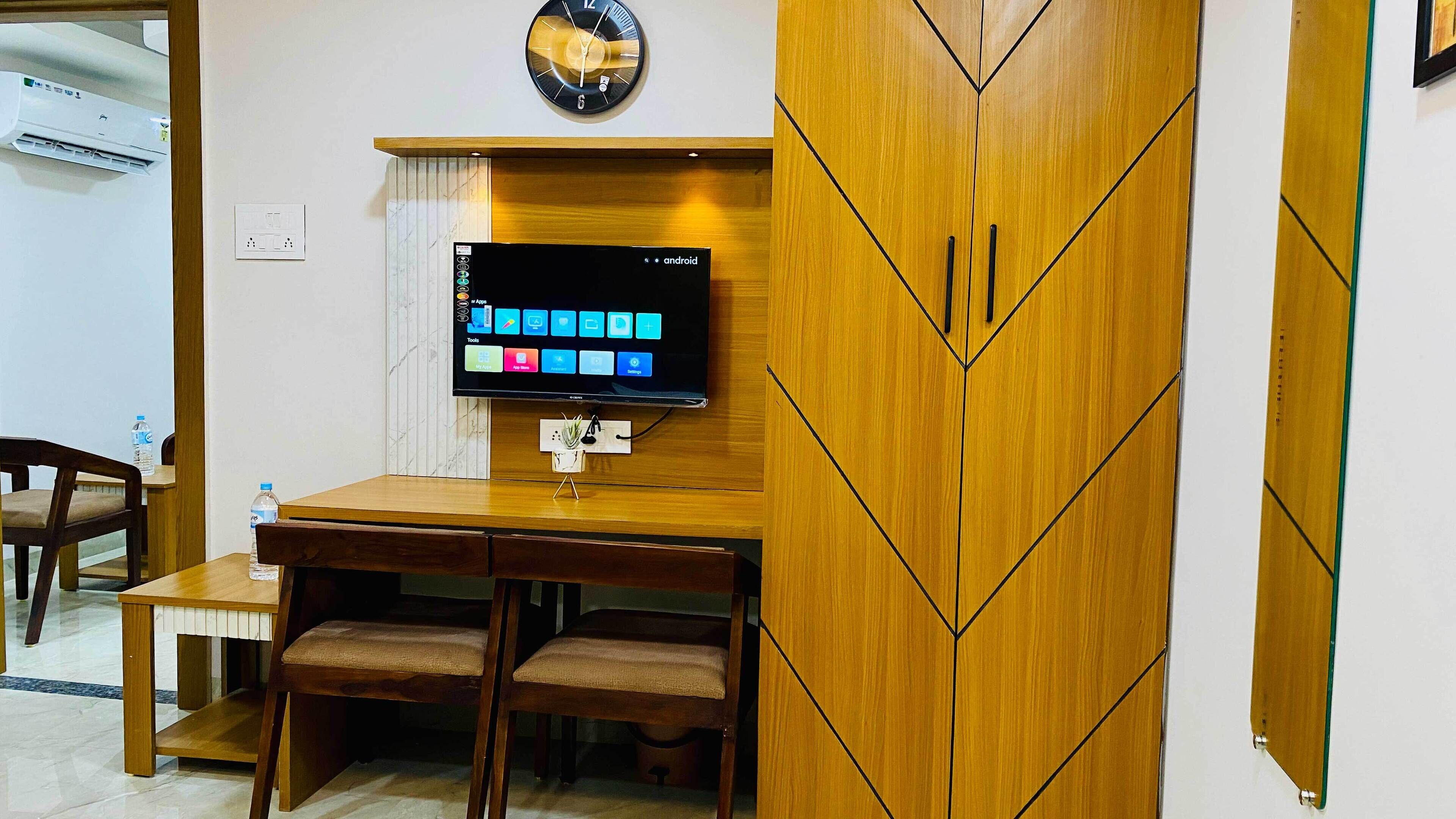 Habitación de hotel con un escritorio de madera, dos sillas, un televisor de pantalla plana en la pared y un reloj encima del televisor.