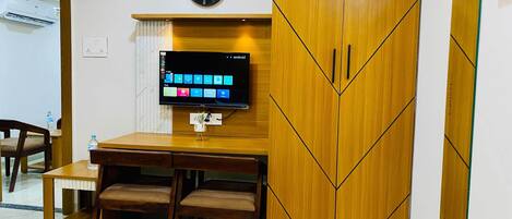 Habitación de hotel con un escritorio de madera, dos sillas, un televisor de pantalla plana en la pared y un reloj encima del televisor.