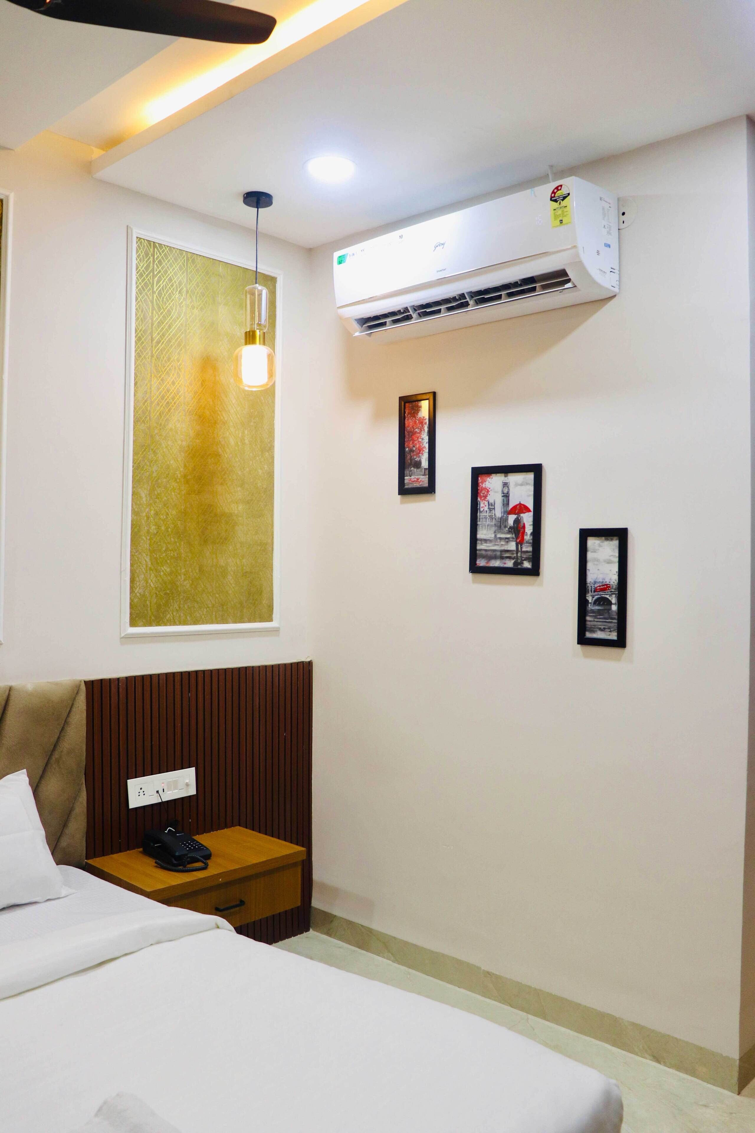A hotel room with a bed, bedside table with a phone, wall-mounted air conditioning unit, and framed pictures on the wall.