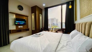 Ein Hotelzimmer mit einem großen Bett, einem an der Wand befestigten Fernseher, einem Fenster mit Blick auf die Stadt und einem kleinen Tisch mit einem Stuhl.