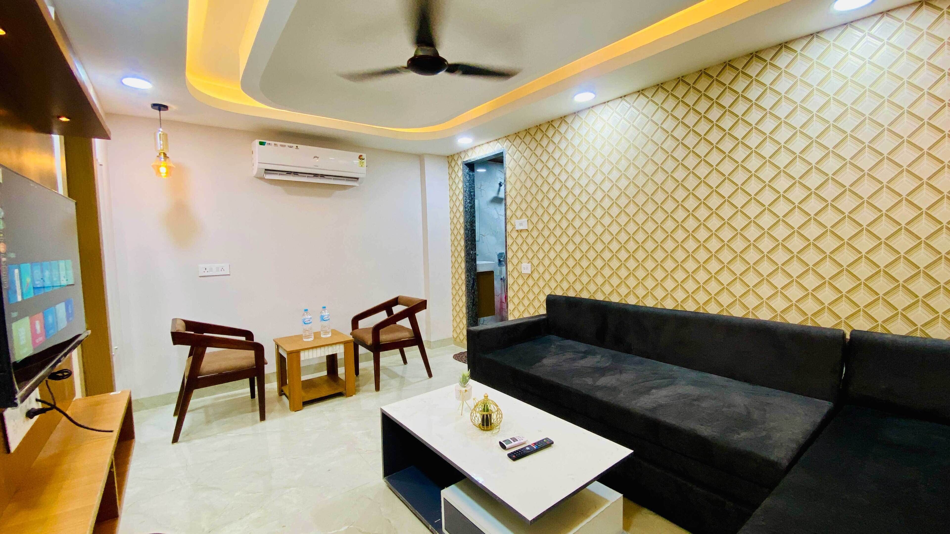 A modern living room with a black sofa, wooden chairs, a small table with two water bottles, a ceiling fan, and an air conditioning unit.