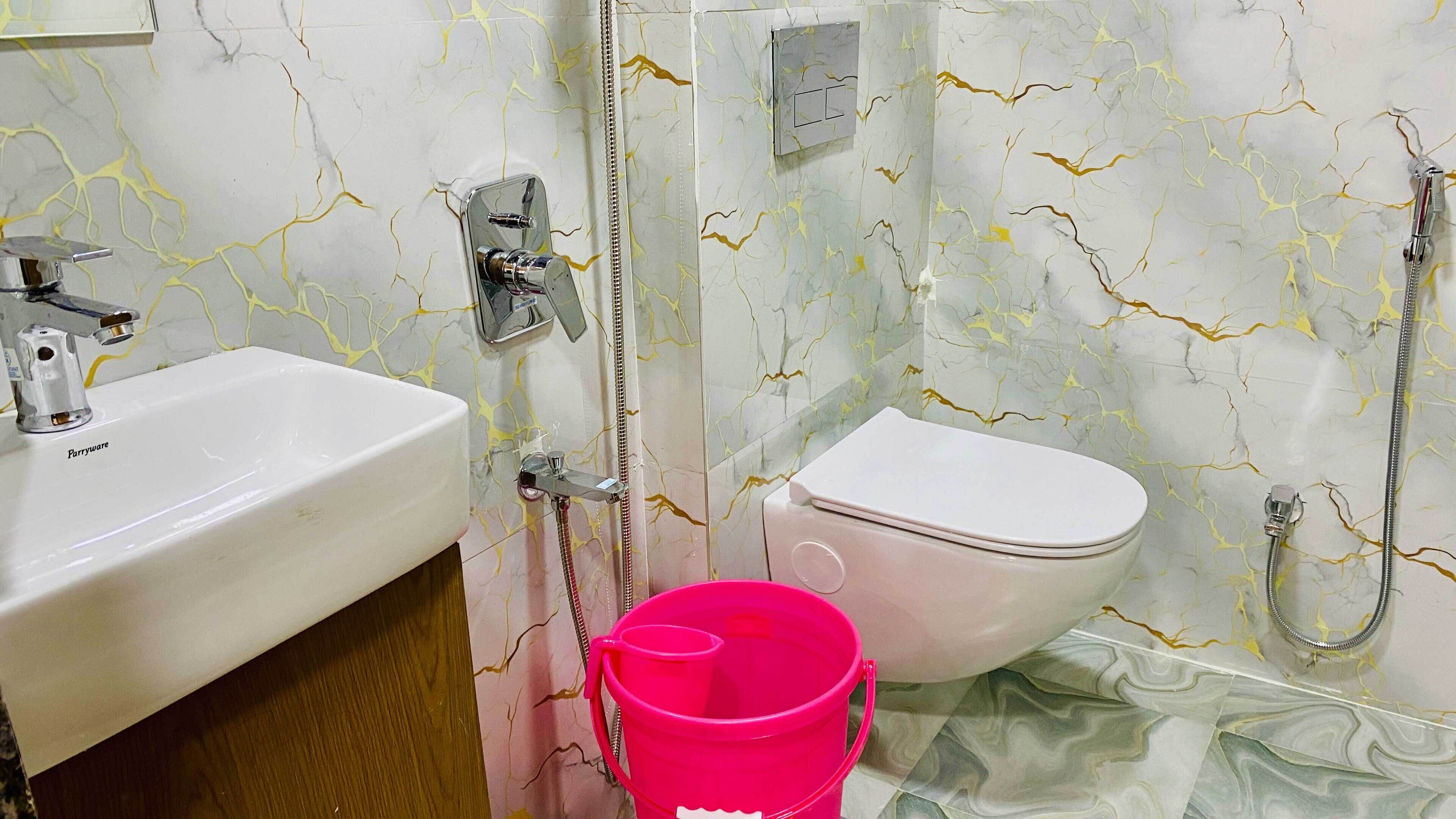 A bathroom with a white sink, a pink bucket, a toilet, and a shower with a hose.