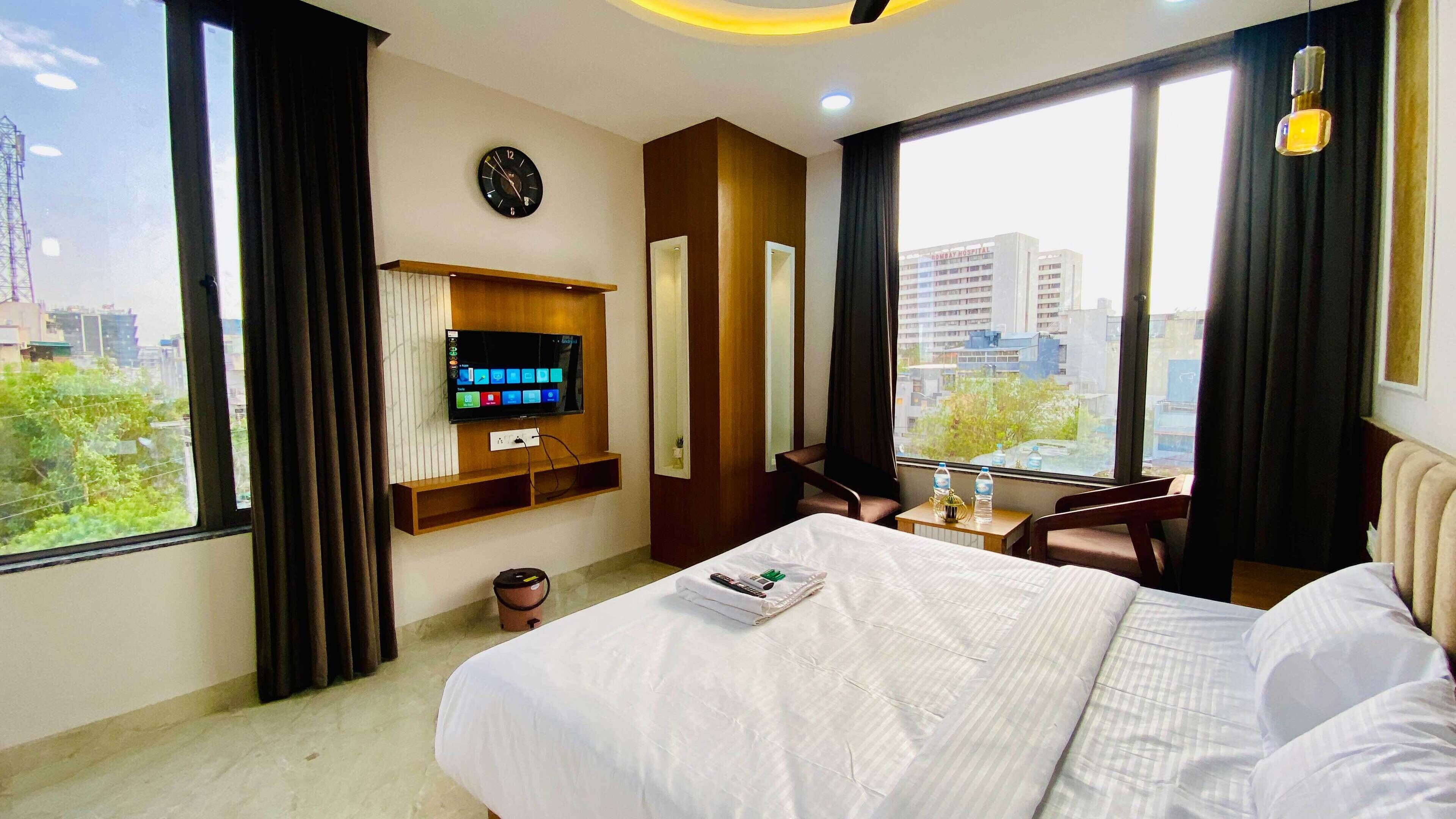 Ein Hotelzimmer mit Bett, Wandfernseher, Schreibtisch mit Stuhl und Blick auf die Stadt durch große Fenster.