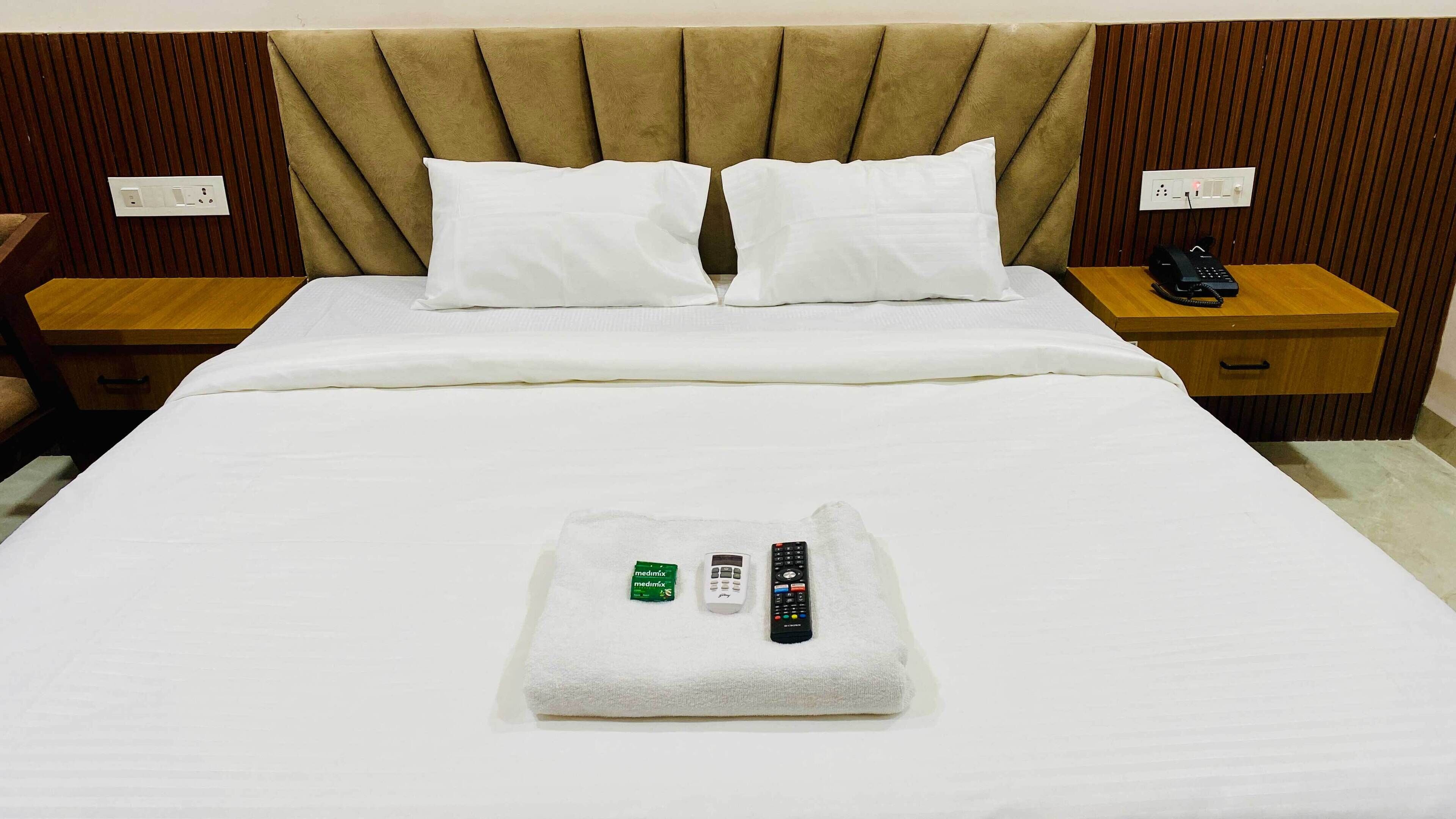 A hotel room with a bed, two pillows, a nightstand with a phone, and a towel with remote controls.