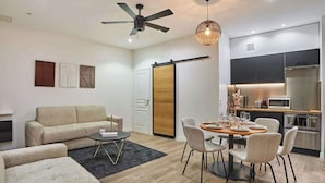 A modern living room with a sofa, a dining table with chairs, a ceiling fan, and a kitchen area with appliances.