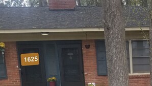 A house with a number sign '1625' on the front door, a small porch with a potted plant, and a tree in the foreground.
