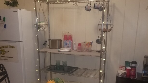 A dining area with a glass table set for two, surrounded by chairs. A metal shelf unit with hanging lights and kitchen items is behind the table.