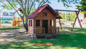 A wooden playhouse with a red roof and a small porch, surrounded by a playground with swings and slides. - Apartamento em Gravatá com Lazer Completo (Gravatá)