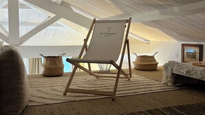 A high-ceilinged room with a wooden ceiling and a large window. A foldable chair with a sign reading "Marie La Valentine" is placed in the center. The room has a bed with a white bedspread and a framed picture on the wall. - House Bormes village La Valentine, 2 levels, near shops, Air-conditioned (Bormes-les-Mimosas)
