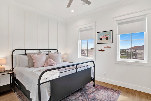 Une chambre à coucher avec un lit en métal noir, des murs blancs et une grande fenêtre donnant sur des maisons.