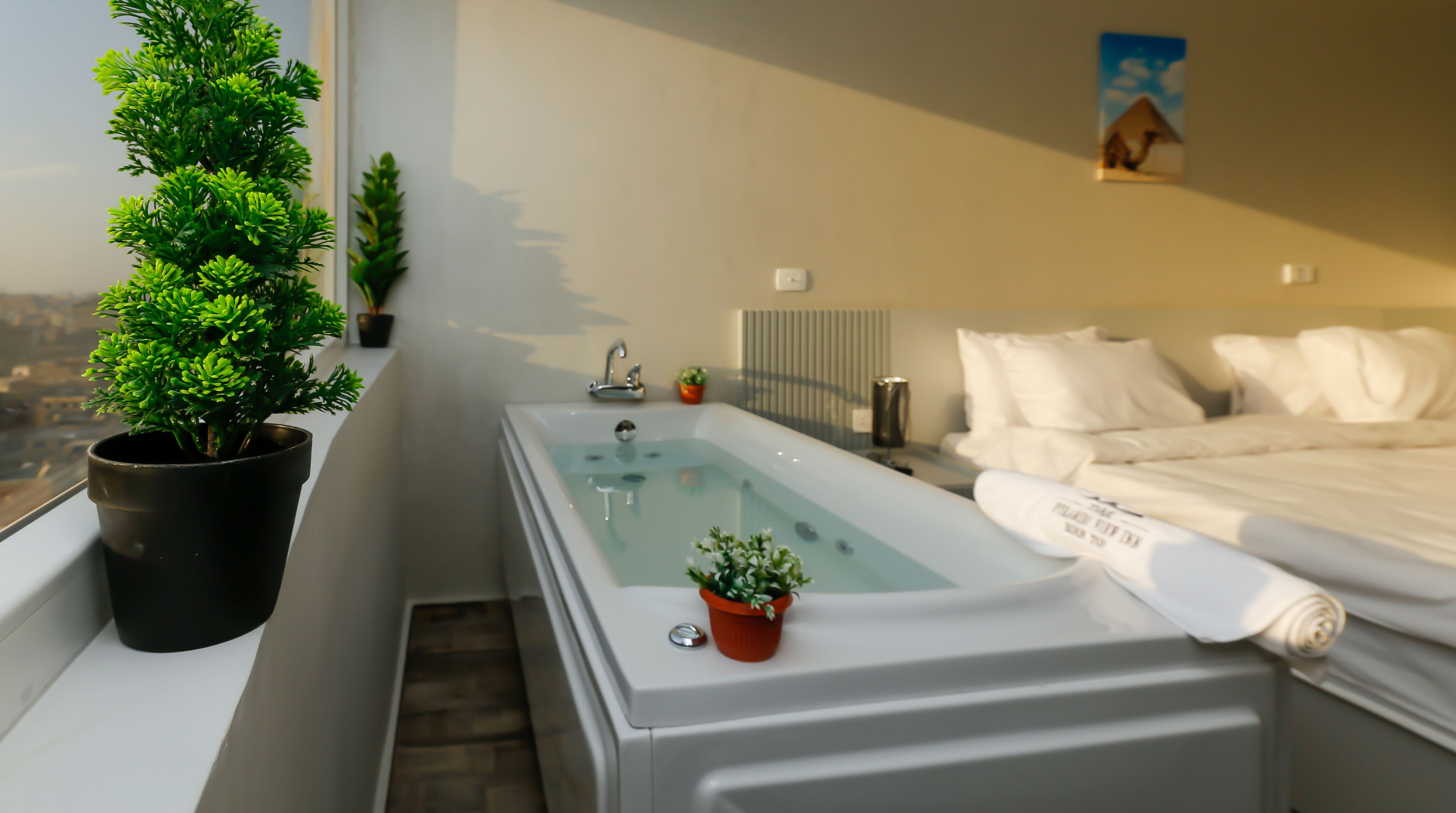 A bedroom with a large bathtub, a bed with white linens, and a potted plant.