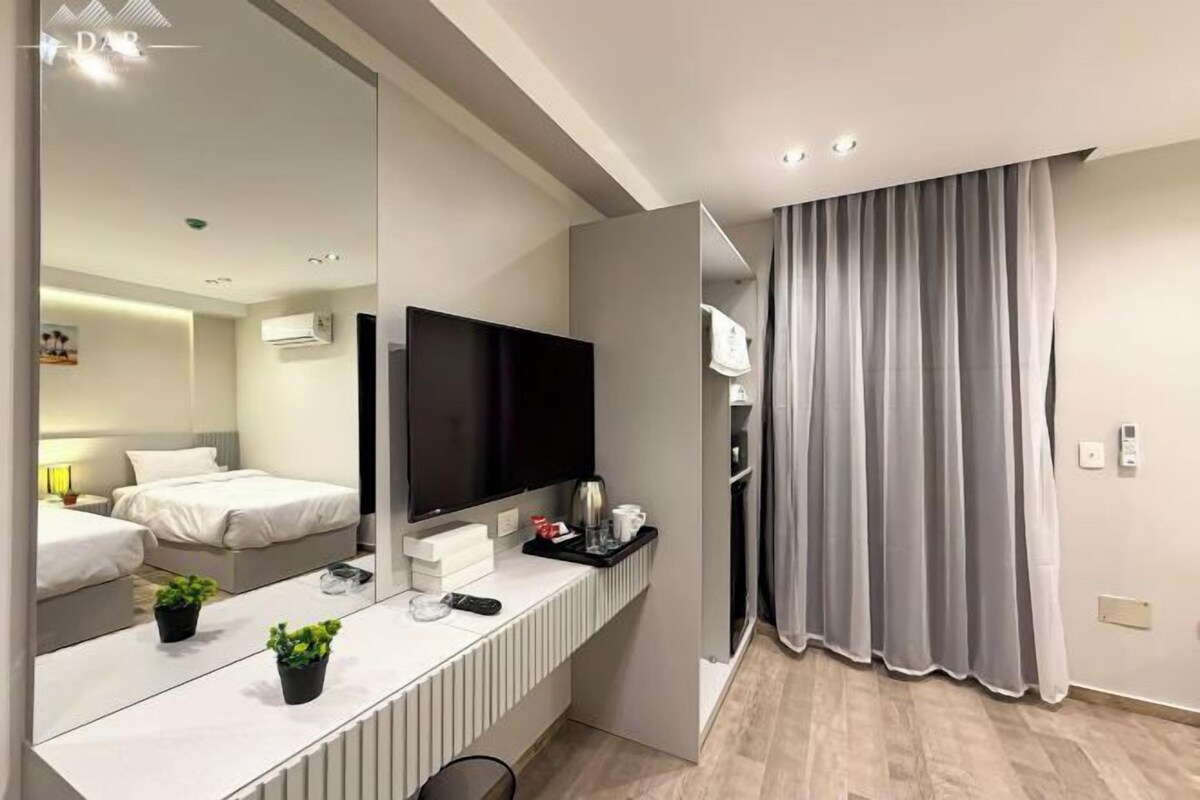 Een moderne hotelkamer met een grote spiegel, een flatscreen televisie, een nachtkastje met afstandsbediening, een kleine plant en een bed met wit beddengoed.