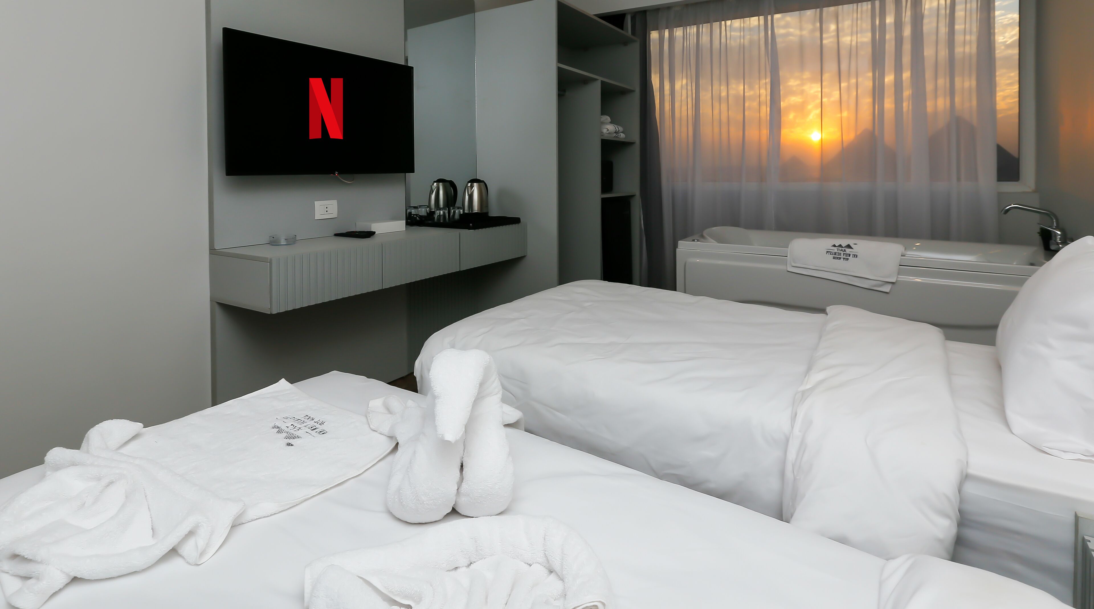 A hotel room with two beds, a TV displaying the Netflix logo, a minibar, and a view of the sunset through sheer curtains.
