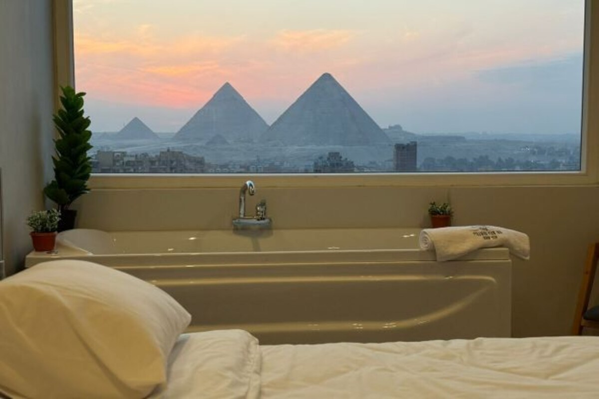 Une chambre avec une baignoire, une fenêtre donnant sur les pyramides et une plante posée sur la table de chevet.