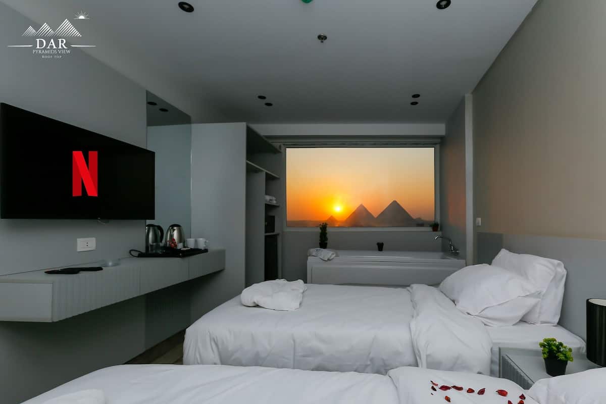 A modern hotel room with a large TV displaying the Netflix logo, a bed with white bedding, a window with a sunset view, and a small plant on the bedside table.