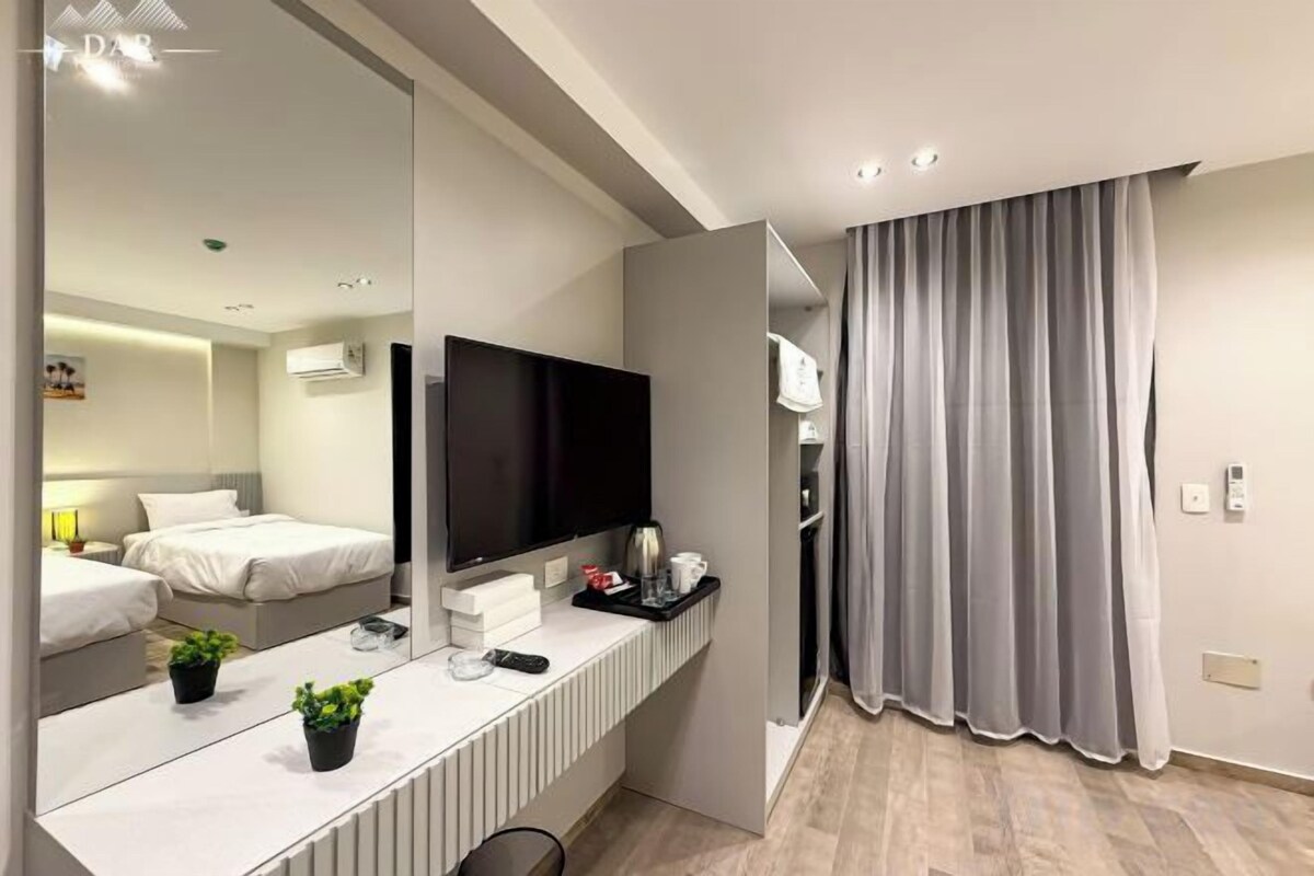 A modern hotel room with a large mirror, a flat-screen TV, a bedside table with a remote control, and a window with curtains.