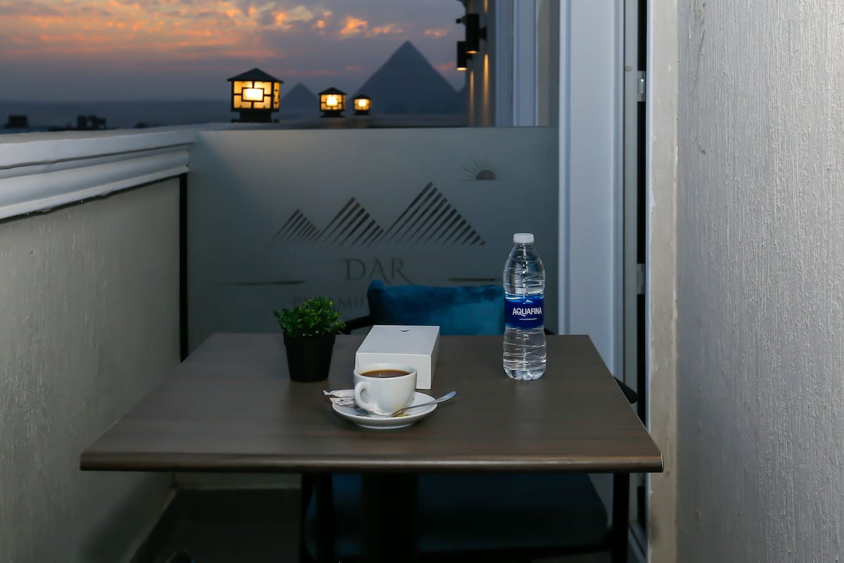 A balcony with a table, a cup of coffee, a water bottle, a small plant, and a view of the pyramids at sunset.