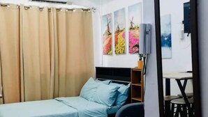 Ein Schlafzimmer mit einem Bett, einem Schreibtisch und einem Sessel. Es gibt ein Fenster mit Vorhängen, eine Wandklimaanlage und ein Gemälde.