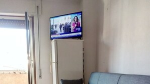 A living room with a grey sofa, a television mounted on the wall, and a tiled floor.