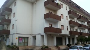 A multi-story residential building with balconies, a front entrance, and a parking area.