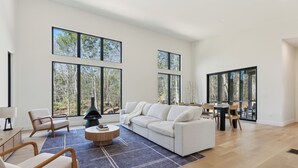 A spacious living room with a large white sofa, a round coffee table, and a dining area with a wooden table and chairs. Large windows provide a view of the outdoors.