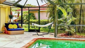 Un espace piscine aménagé avec un hamac, un banc rembourré et une vue sur une maison et une végétation tropicale.