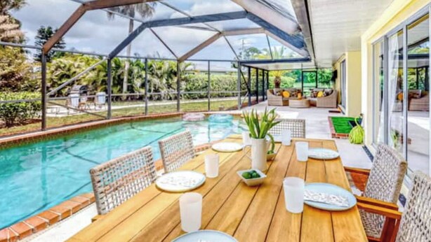 Un espace piscine extérieur abrité, doté d’une table et de chaises en bois, entouré de murs en verre et surmonté d’une structure de toit en métal.