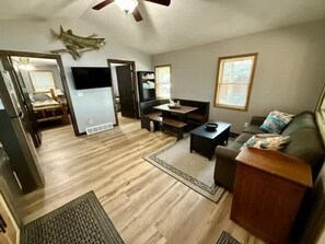 A living room with a sofa, coffee table, and TV. There is a dining area with a table and chairs. The room has hardwood flooring and a ceiling fan.