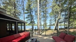 A deck with red outdoor furniture, a metal railing, and a view of a lake surrounded by trees. - Musky Hideout on Lost Land Lake (Hayward)