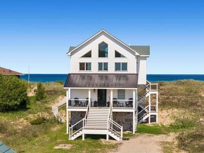 A two-story beachfront house with a balcony, a stone foundation, and a view of the ocean.