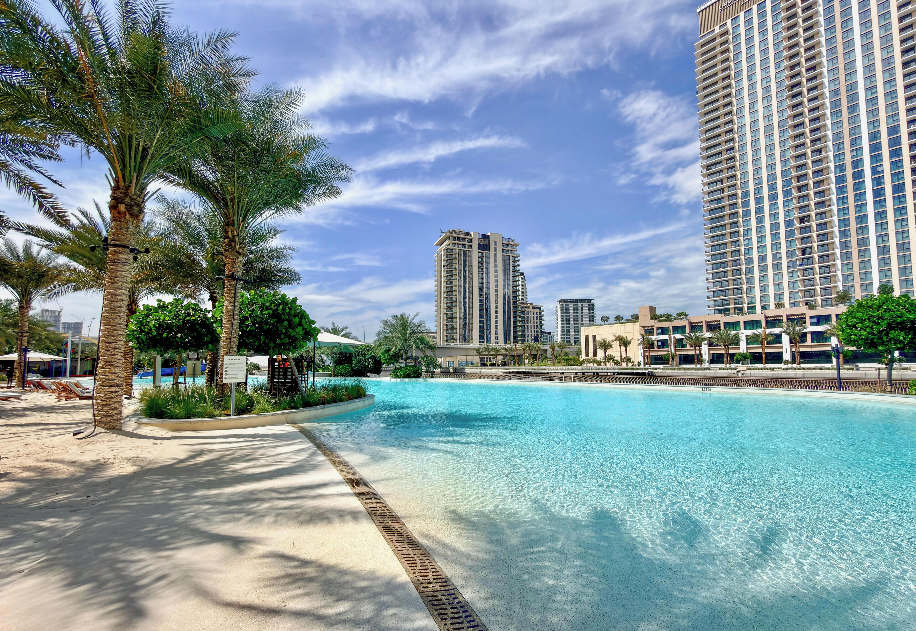 A swimming pool with a sandy edge, surrounded by palm trees and modern high-rise buildings in the background.