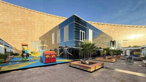 Een speeltuin met glijbanen en klimconstructies, omringd door een gebouw met grote ramen en een betegeld plein.