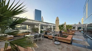 Een terras op het dak met buitenmeubilair, een zwembad en uitzicht op een modern gebouw en een klokkentoren.