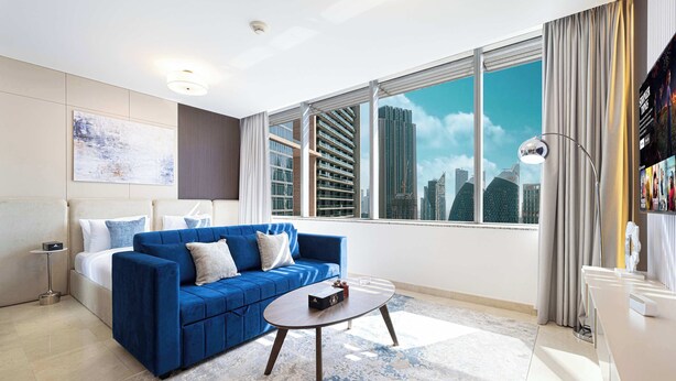 Een moderne hotelkamer met een blauwe bank, een ronde salontafel, een bed, een nachtkastje en grote ramen met uitzicht op de stad.